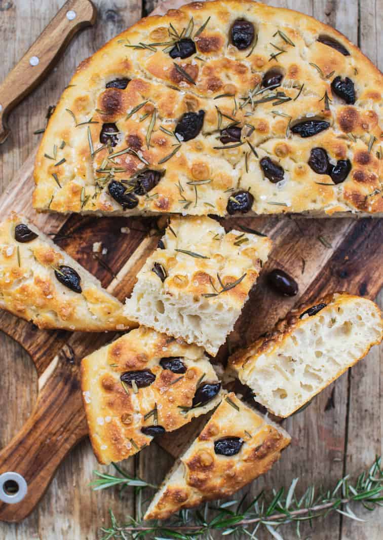 a round focaccia with olives and rosemary on a wooden board with some pieces cut off