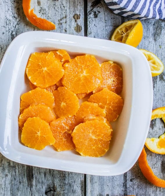 oranges macerated with lemon juice in a white tray sitting on grey wood with squeezed lemons and orange peel on the wood