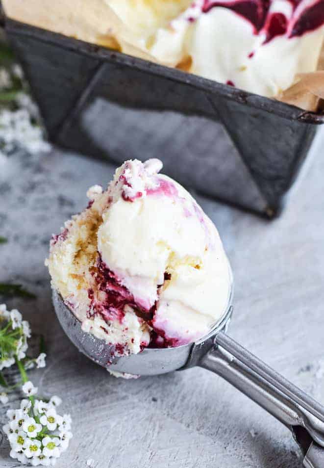 ice cream scoop filled with a scoop of black berry ripple ice cream sitting on a bench. loaf tin behind ice cream scoop and and small white flower to the side