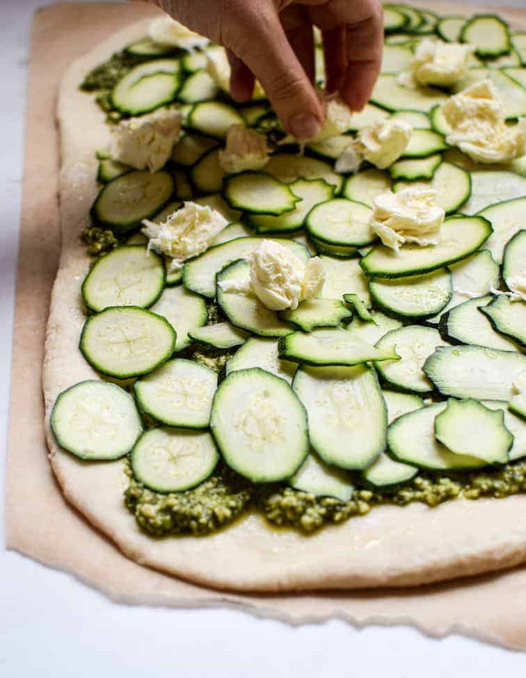 a hand dropping mozzarella pieces over slices of zucchini layered on a pizza base