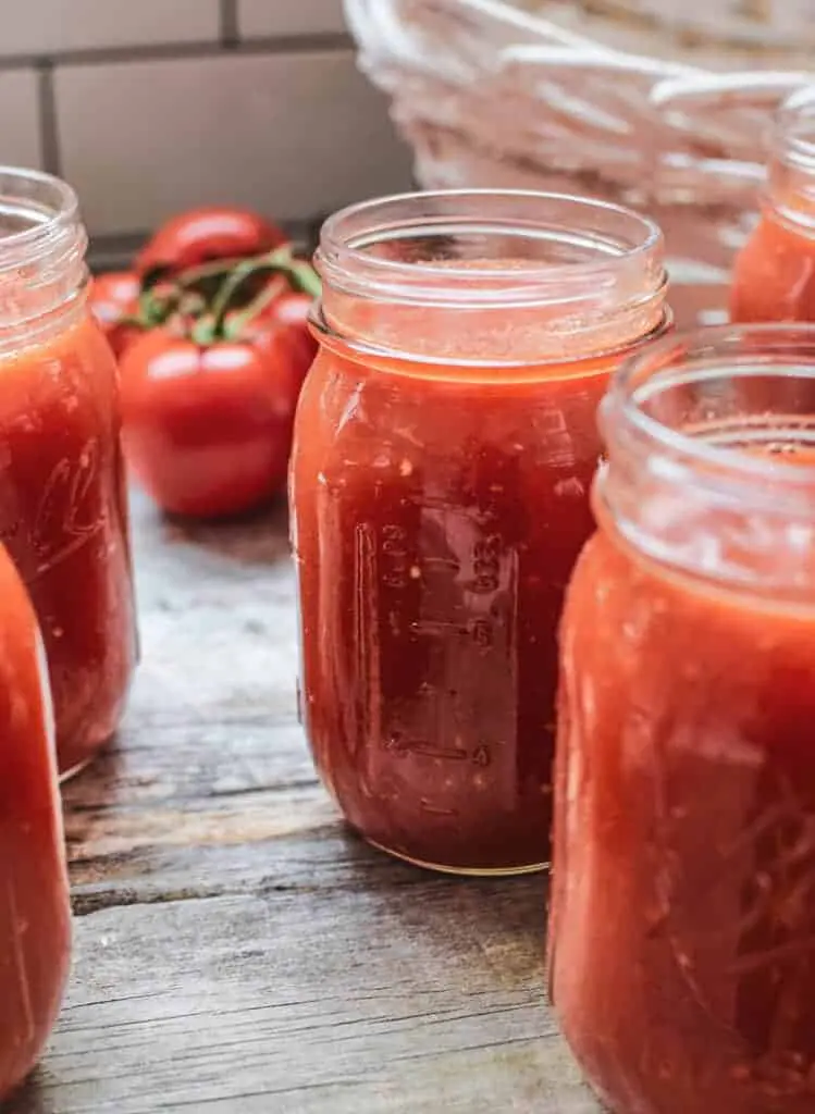 Close up of 5 jars without lids on a wooden table close filled with tomato passata. A white basket in the top right out of focus and 4 on the vine red tomatoes in the background out of focus.