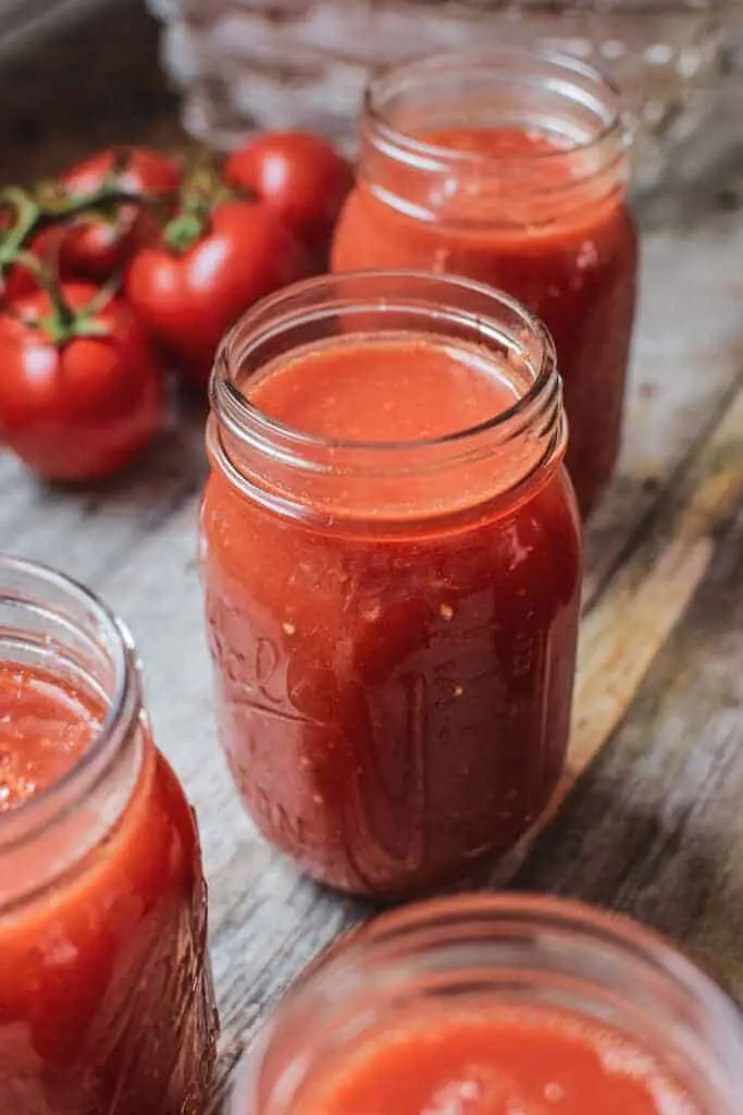Close up of 4 jars without lids on a wooden table close filled with tomato passata. 5 on the vine red tomatoes in the top left out of focus.