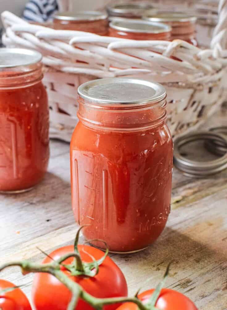 Close up of 2 jars of tomato passata with lids on a wooden table and 3 on the vine tomatoes at the bottom of the image. Behind the jars at the top is a white basket with 4 other sealed jars inside.