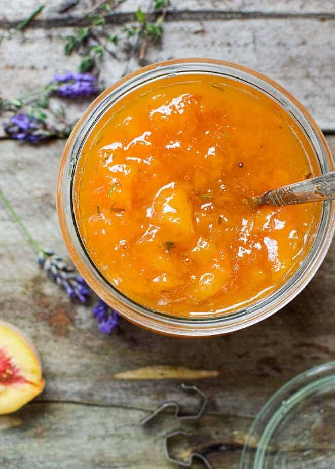 Birds eye view of a jar filled with orange peach preserve with thyme leaves and a spoon handle poking out the top of the jar. The jar is on top of a wooden background with lavender flowers scattered around a half peach and a jar lid.