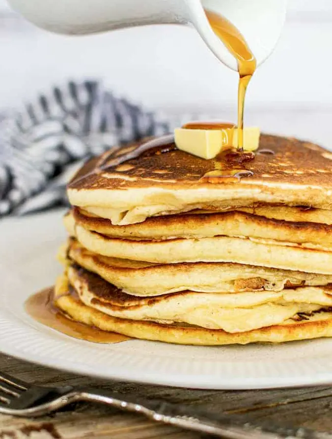 a stack of pancakes with a knob of butter and maple syrup being poured over