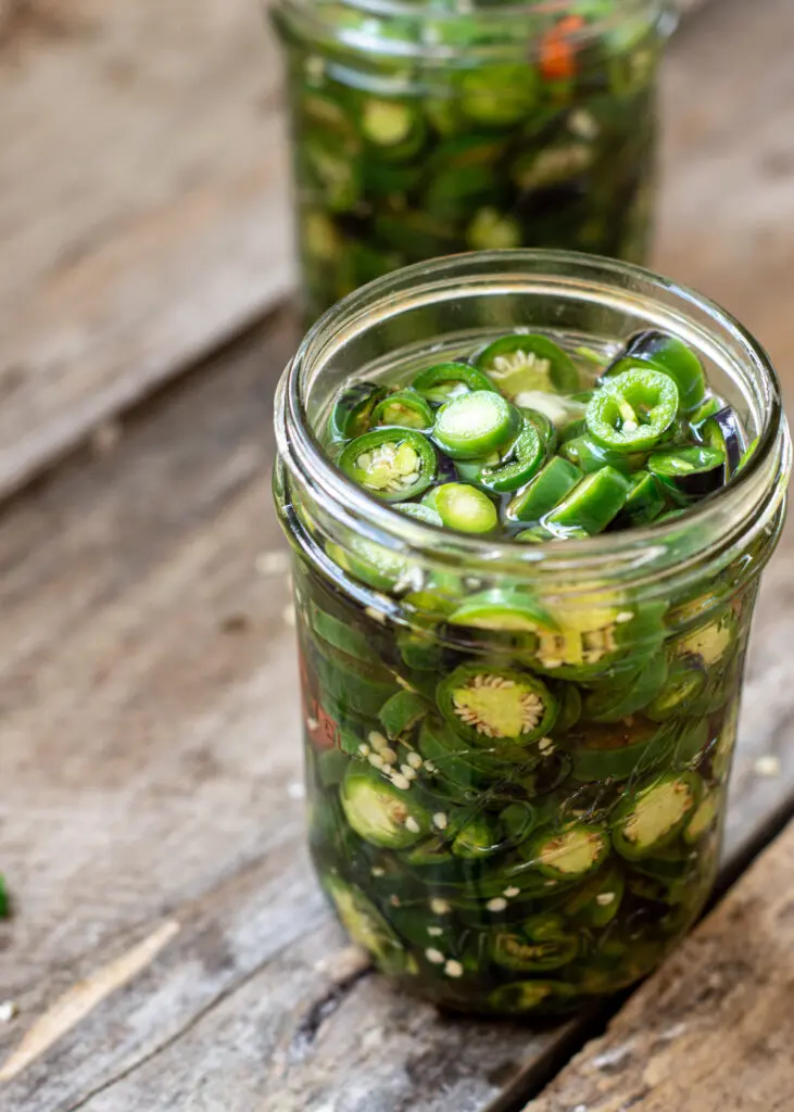 a glass jar of bright green jalapeño pepper slices on a wooden board. Another jar is in the background